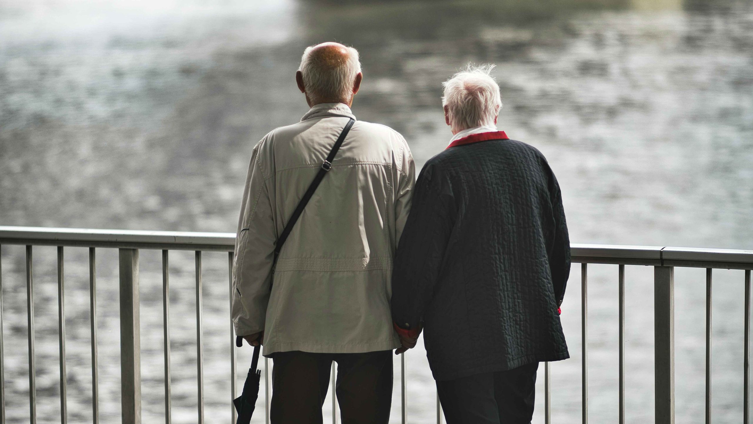 An older couple standing together and looking at a river from the shore.